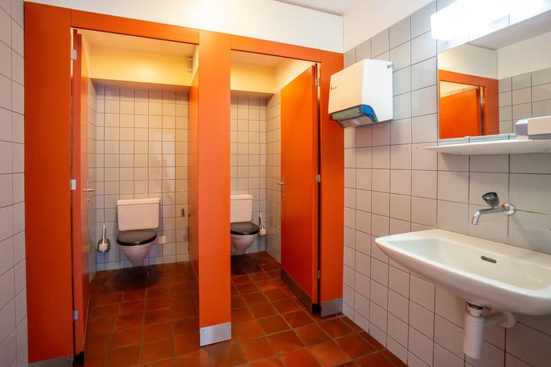Two toilets, sink, mirror, orange walls, tiled floor