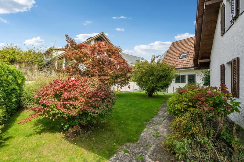 Backyard garden with red and green plants, manicured lawn, wooden path, houses in background