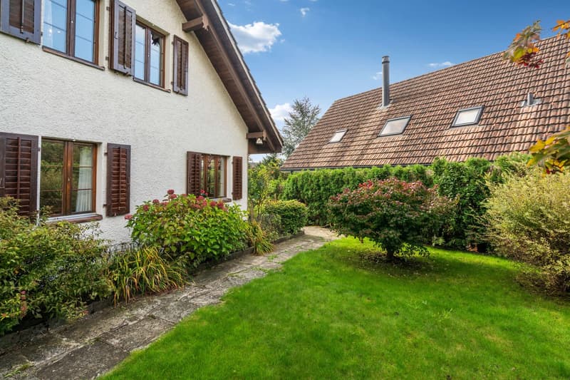 Two houses with brown roofs, windows with shutters, green grass, bushes, and plants