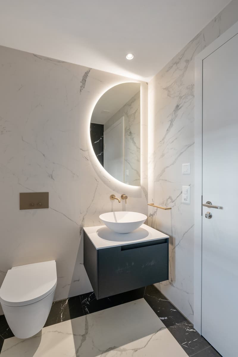 modern bathroom with marble tile walls, round mirror, white porcelain sink, brass faucet, modern toilet, marble tile flooring