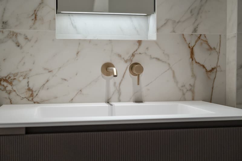 modern bathroom with marble walls and white countertop, brass faucets, mirror with light