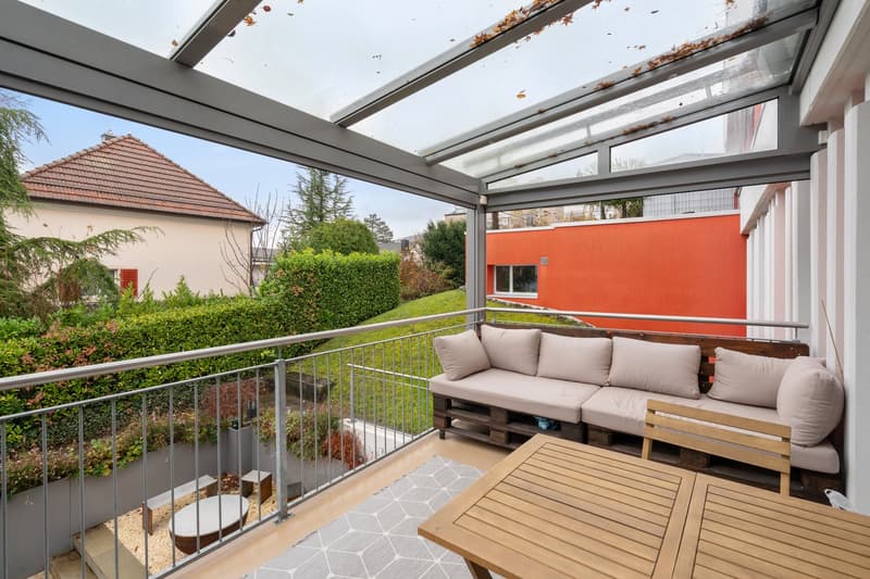 Terrace with wooden sofa, glass roof, metal railing, wooden table, rug, plants, and a building in the background.