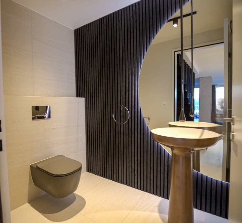 modern bathroom with black wall, white tiles, a toilet, two sinks, a circular mirror