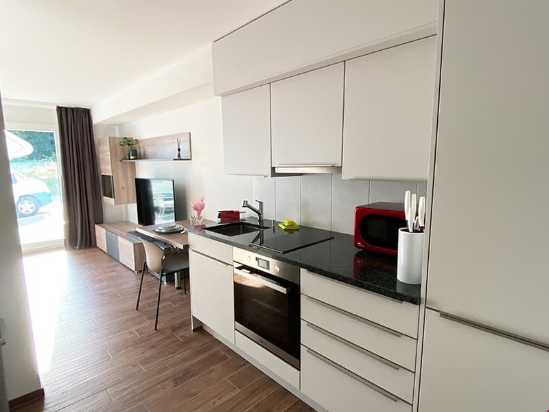 modern kitchen with wooden floors, black countertops, white cabinets, large windows, TV, desk, chairs, built-in oven, microwave, sink, and faucet