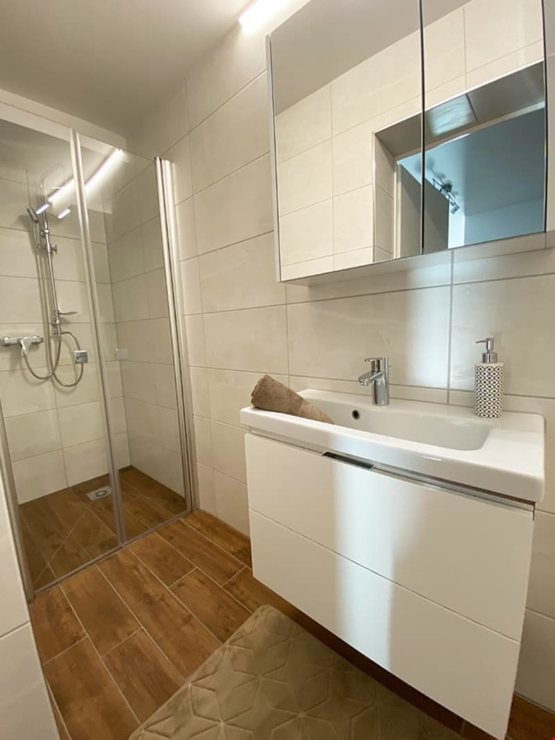 White bathroom with tiled flooring, walk-in shower with shower head, white sink with a faucet, and a mirror.