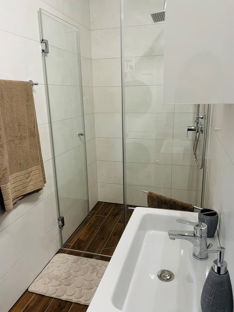 Bathroom with white tile walls, glass shower enclosure, wooden floor tiles, sink with faucet, and a towel rack.