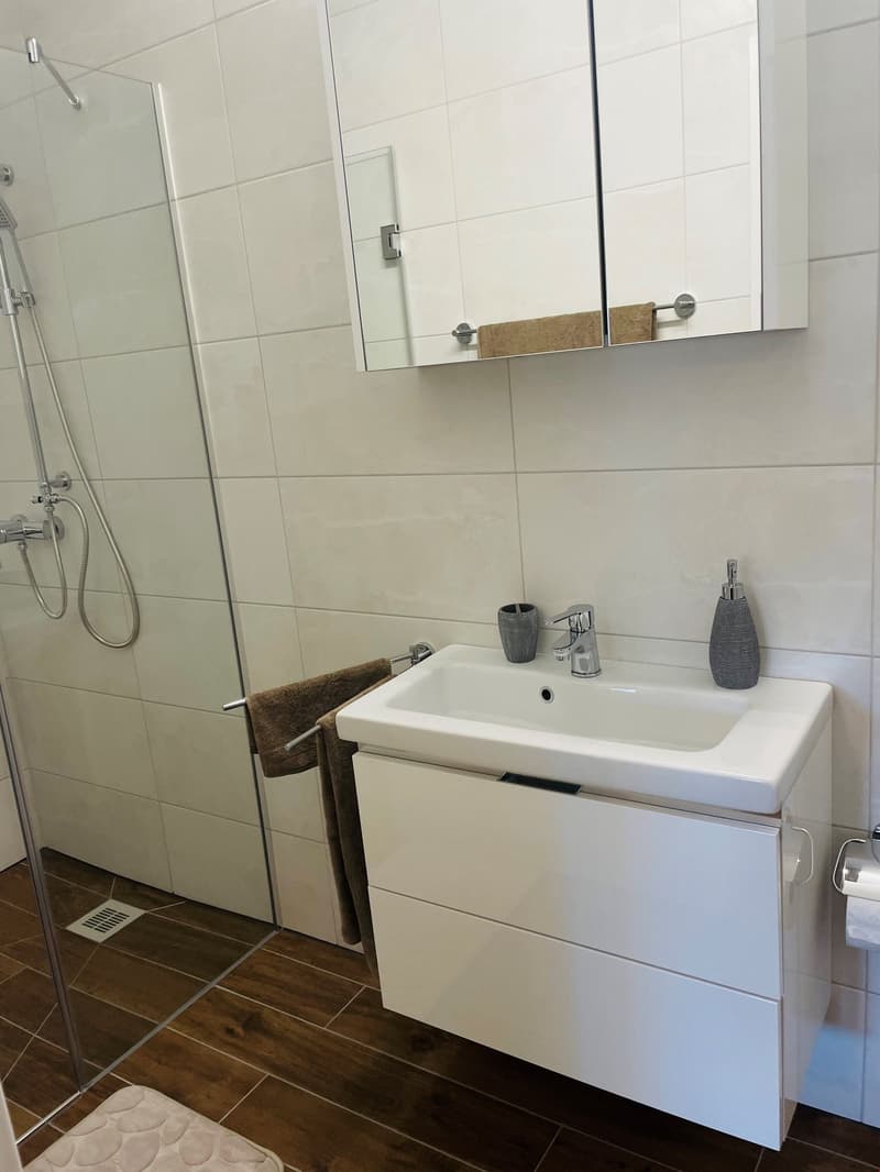 Bathroom with white tile walls, vanity with sink, mirror cabinet, shower enclosure, and wooden floor tiles.