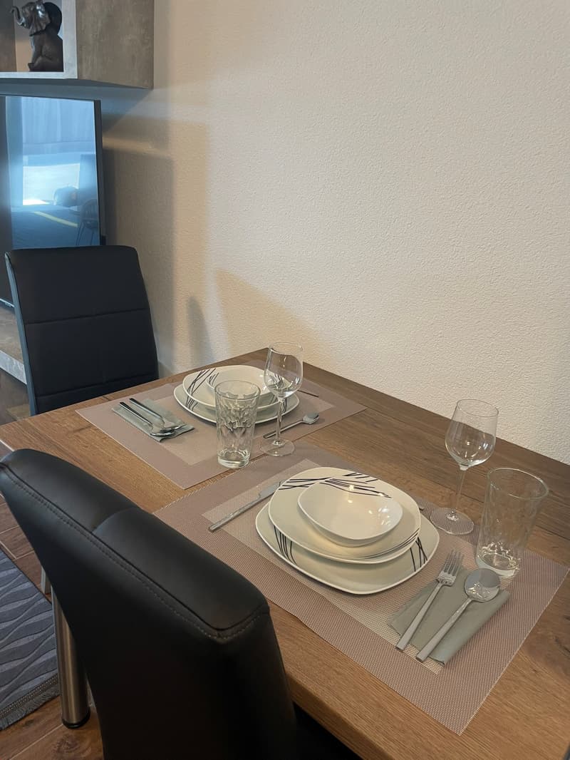 The image shows a dining table set with plates, glasses, and utensils. The table has a wooden surface and is surrounded by black leather chairs. There is a framed artwork on the wall above the table.