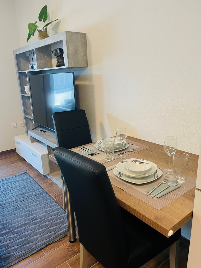 The image shows a dining area with a wooden table set for a meal, including plates, glasses, and utensils. There is a black leather chair at the table. Behind the table is a shelving unit with a plant on top and what appears to be a dishwasher visible in