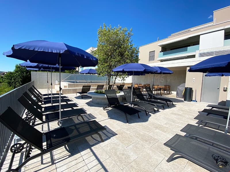 The image shows an outdoor patio area with several lounge chairs and umbrellas. The patio has a tiled floor and is surrounded by a building with balconies. There is a tree in the center of the patio, providing some shade.