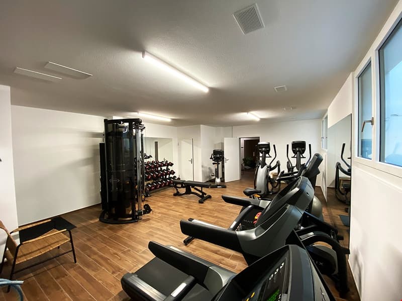 Large open space with hardwood flooring, various exercise equipment including weight machines, treadmills, and other cardio machines, well-lit with overhead lighting