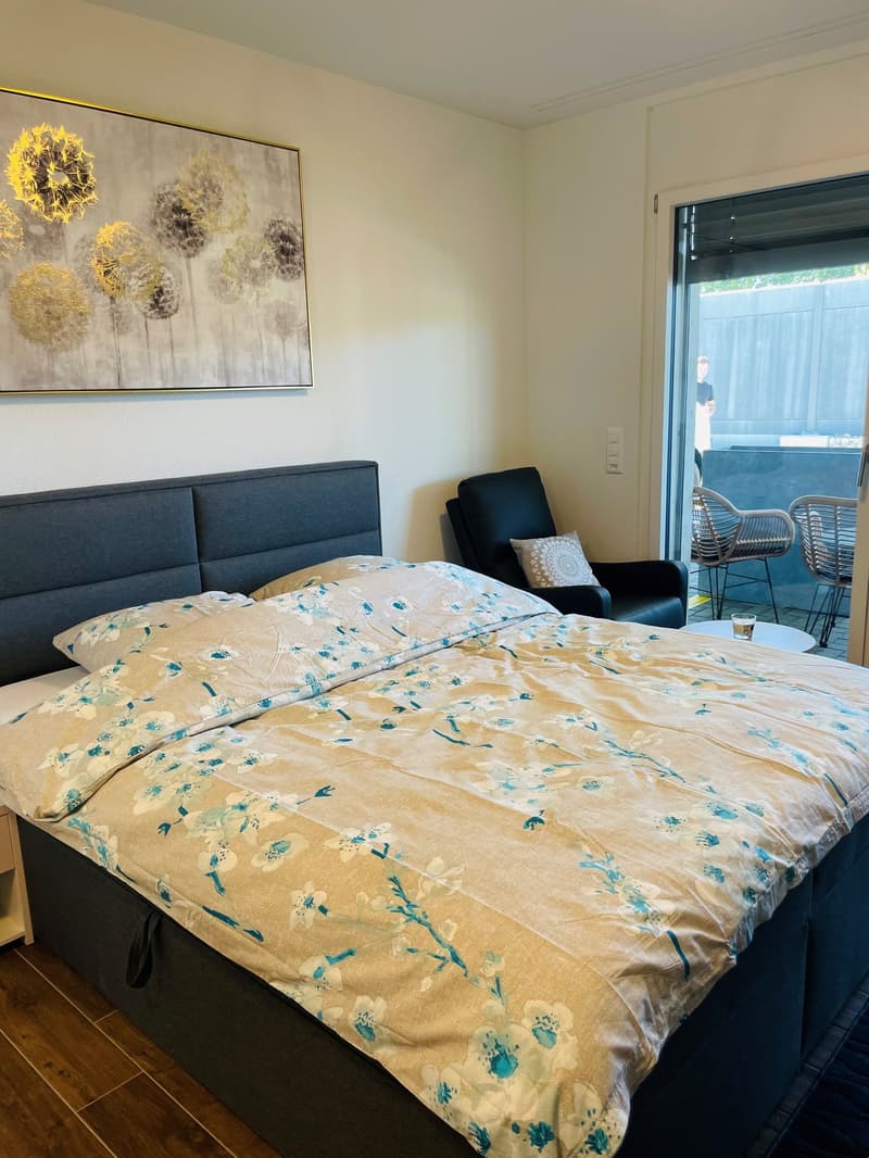 Spacious bedroom with a large bed, floral bedding, and a framed artwork on the wall. There is a sliding glass door leading to a balcony or terrace area.