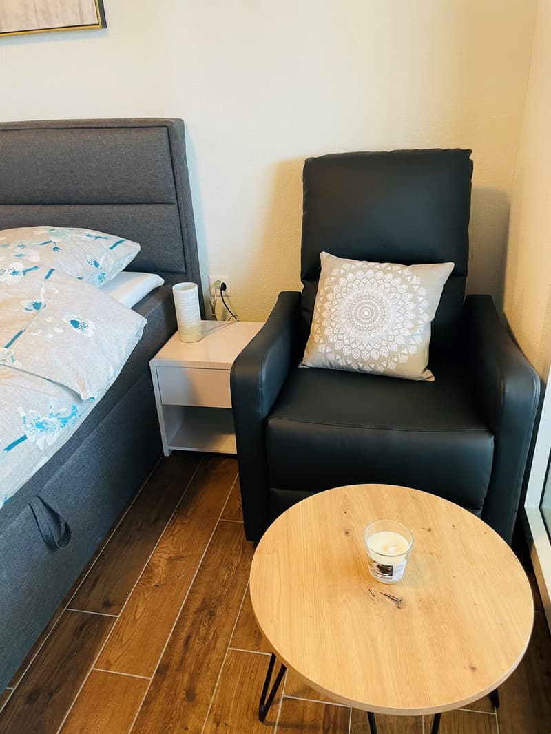 Bedroom with a grey upholstered headboard, a black leather armchair, a wooden side table, and a round wooden coffee table. The room has hardwood floors and a decorative pillow on the armchair.