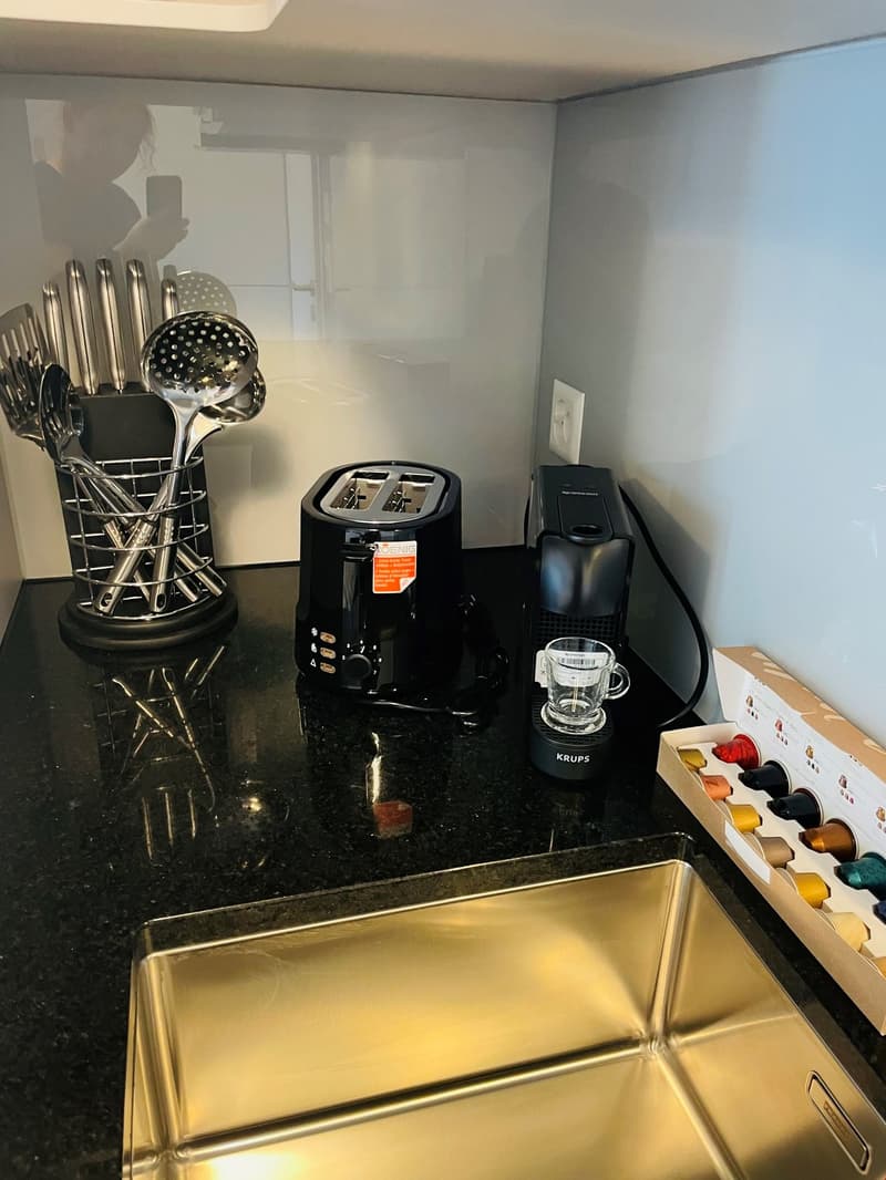 The image shows a kitchen counter with various kitchen appliances and utensils. There is a Krups coffee maker, a toaster, and a dishwasher visible on the counter. A rack of kitchen utensils is also present. The counter appears to be made of black granite