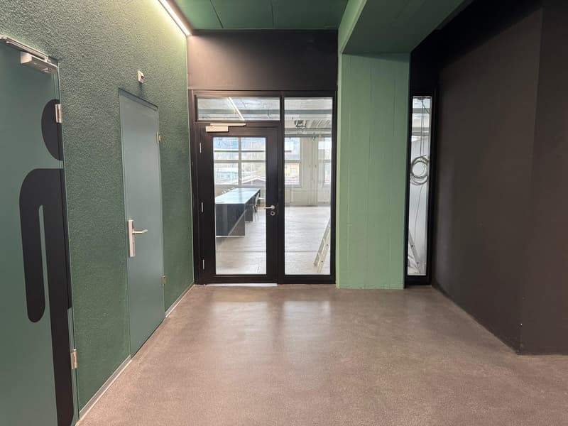 The image shows an interior office space with green walls, a black door with a glass panel, and a concrete floor. The space appears to be a commercial or industrial setting, possibly an office or workspace.
