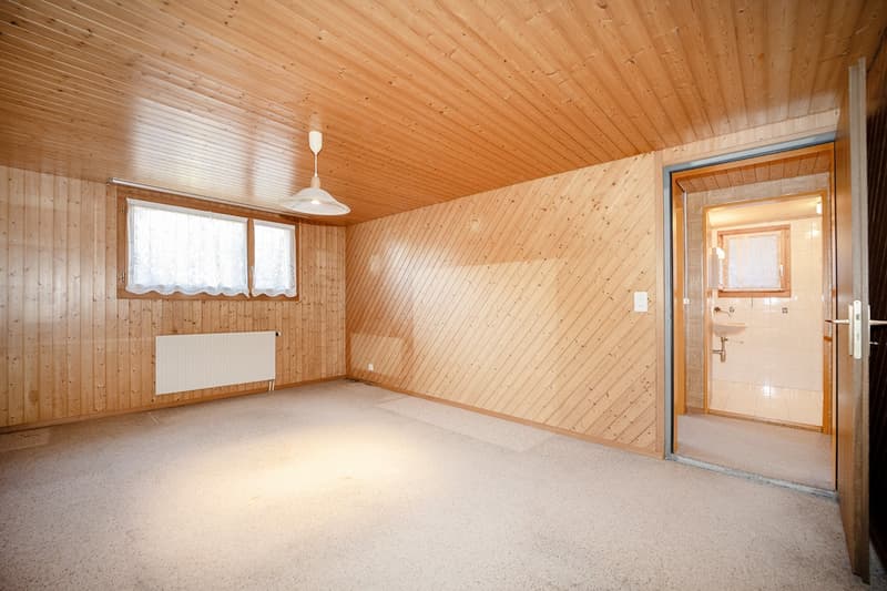 A room with wooden walls, wooden ceiling, windows with white curtains, radiator, and a door leading to a bathroom.