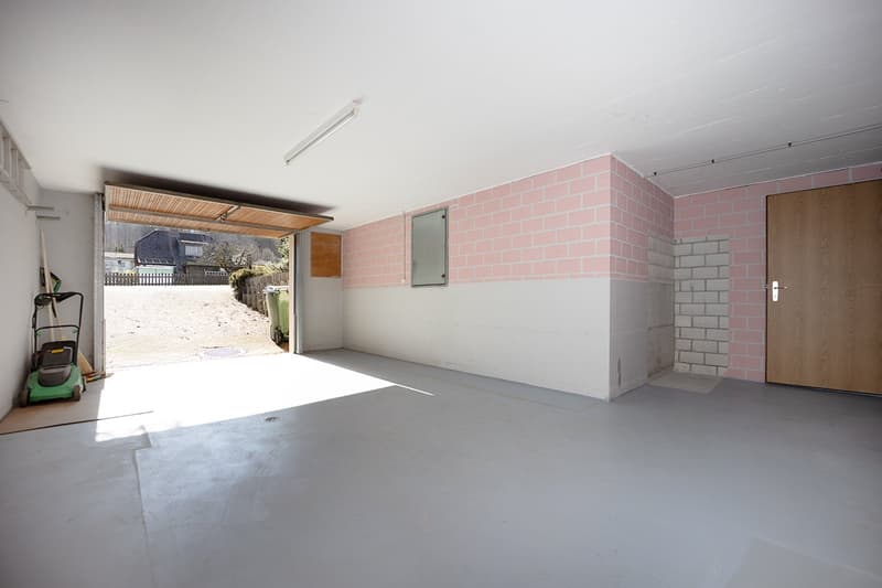Garage with pink brick walls, open door, wooden floor, a small broom, and a lawn mower.