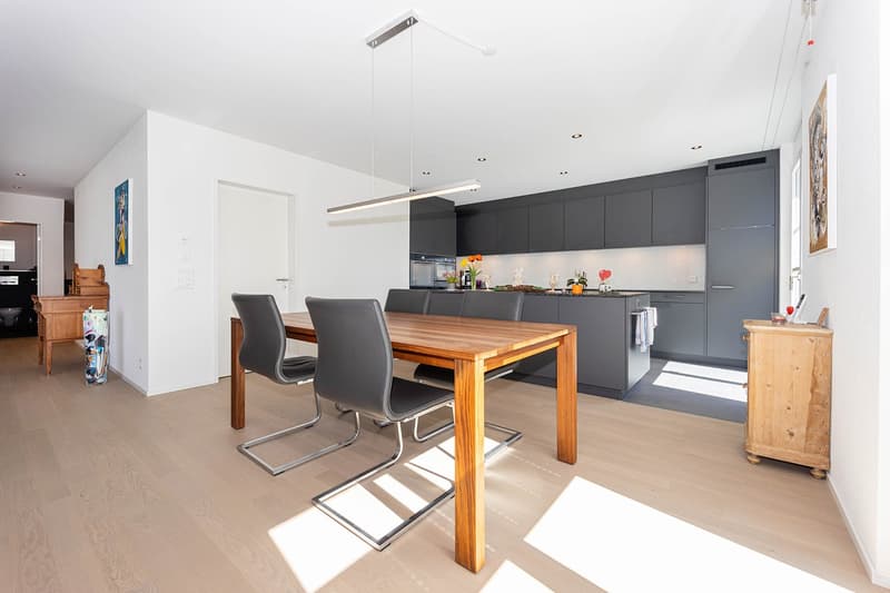 Dining room with wooden table, gray chairs, modern kitchen with gray cabinets, wooden floor, white walls, pendant light