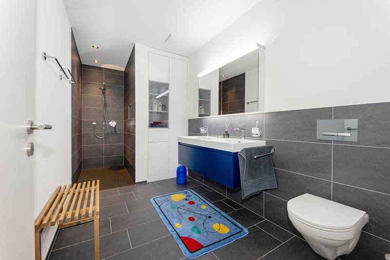 modern bathroom with shower, double sink, toilet, wooden bench, rug, white and gray tiles