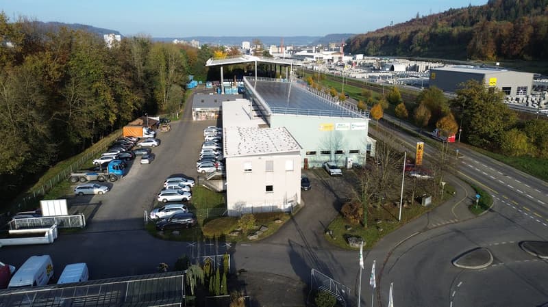 An industrial facility with a large parking area with multiple vehicles parked. A warehouse with a solar panel roof is situated on the property. Several buildings and trees are in the distance.