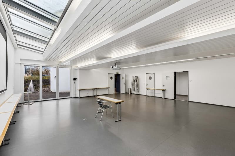 Empty room with white walls, gray floor, glass doors, projector, tables, chairs, and benches