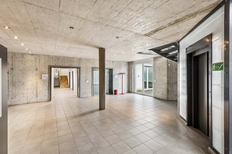 Empty hallway, concrete walls, tiled floor, two elevators, staircase, glass doors, emergency exit sign