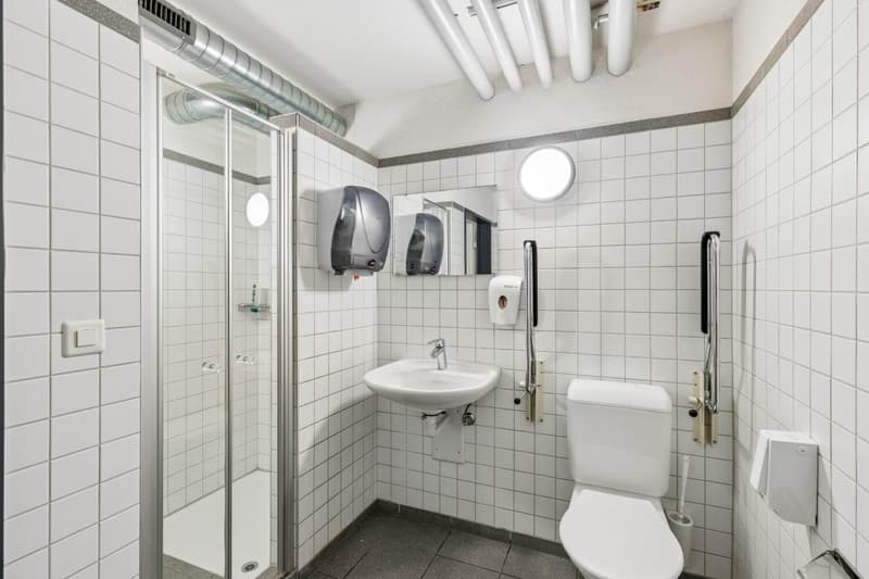 white tiled walls, white tiled floor, shower, sink, toilet, hand dryers, mirror, grab bars
