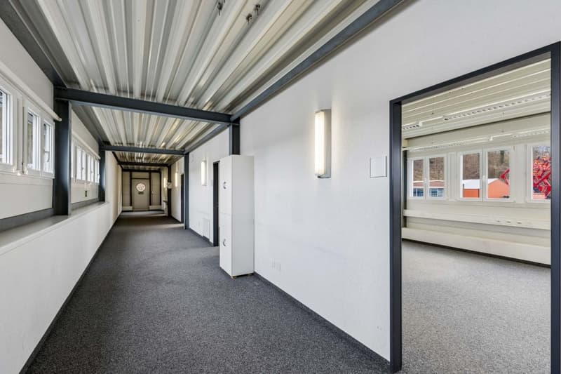 Empty hallway with carpeted floor, white walls, metal ceiling, windows, and wall-mounted lights.