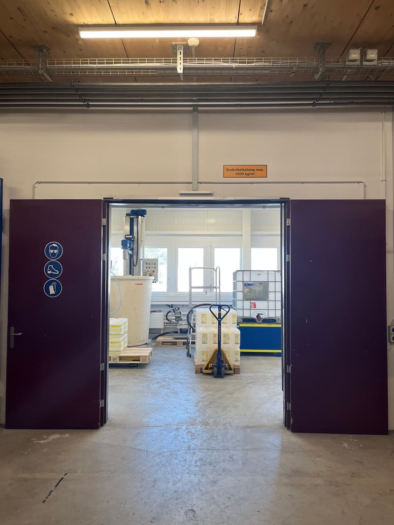 The image shows the interior of a warehouse or storage facility. It has a large open space with a high ceiling and exposed beams. There is a pallet jack and other equipment visible in the background, indicating this is a functional storage or distribution