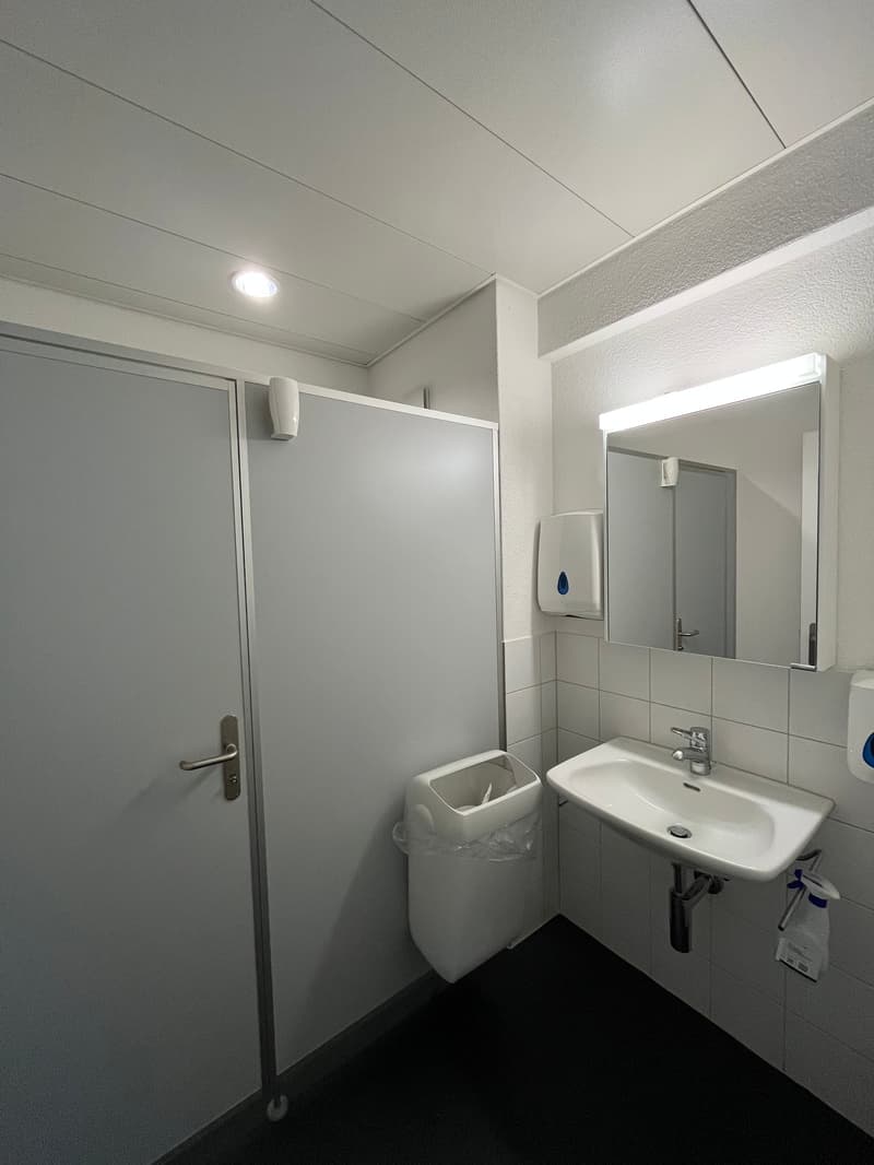 Bathroom with white tile walls, white sink, faucet, mirror with wall mount, trash can, tissue dispenser, under-counter hand dryer, door with silver handle, and white ceiling.