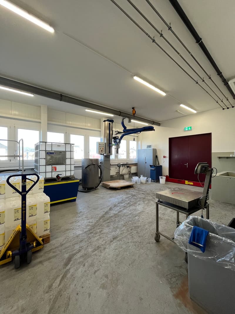 Industrial setting, large floor space, heavy machinery, large steel containers, metal shelves, several large windows, fluorescent ceiling lights, trolley with plastic-wrapped boxes, gray trash bin.