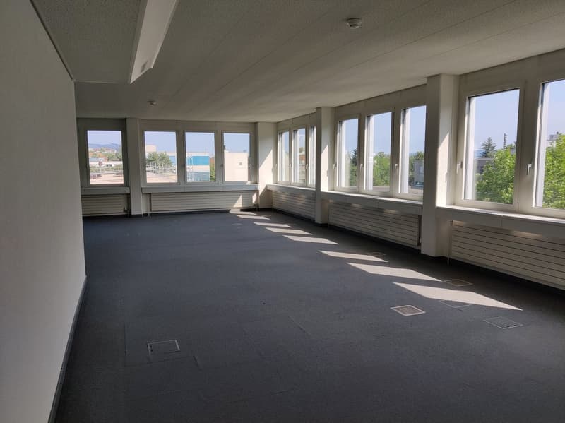 Large open office space with floor-to-ceiling windows, gray carpeted flooring, and white walls. The room appears to be empty and ready for use.