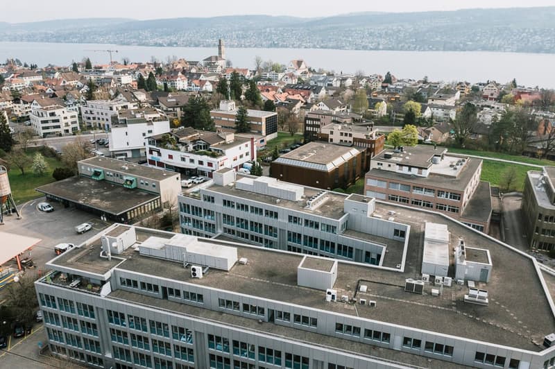 Aerial view of a city with a mix of modern and historic buildings, surrounded by hills and a body of water in the background.