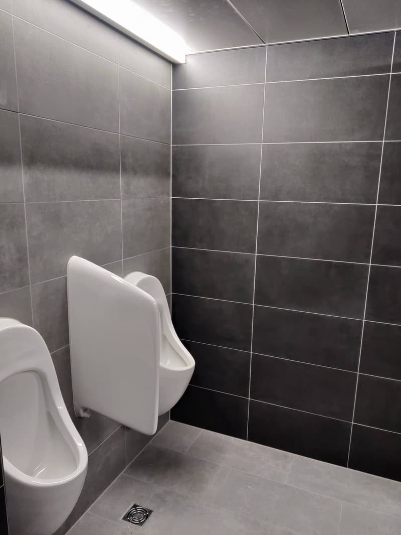 The image shows a bathroom with gray tile walls and floor. There is a white toilet and a drain on the floor.