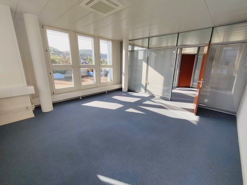 Large open office space with floor-to-ceiling windows, providing a bright and airy atmosphere. The room features a gray carpeted floor, white walls, and a suspended ceiling with air conditioning vents. The space appears to be suitable for use as an office