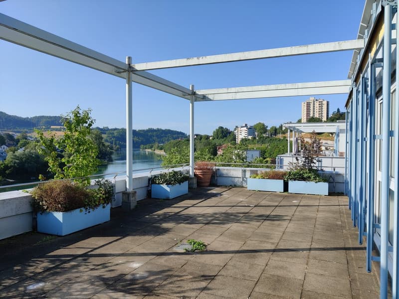 The image shows an outdoor terrace or balcony area with a view of the surrounding landscape. The terrace has a concrete floor and is covered by a metal frame structure. There are several potted plants and flowers placed around the terrace, adding greenery