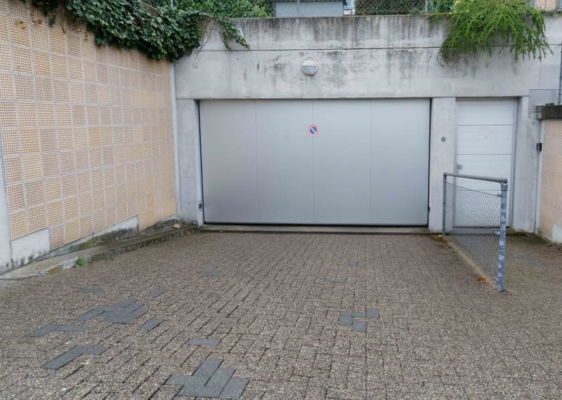 A concrete garage with two doors, one large and one small, and a small paved area in front.