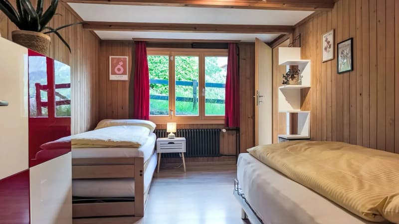 Cozy bedroom with wooden walls and ceiling, large windows overlooking a garden, red curtains, two beds, a nightstand, and a shelving unit