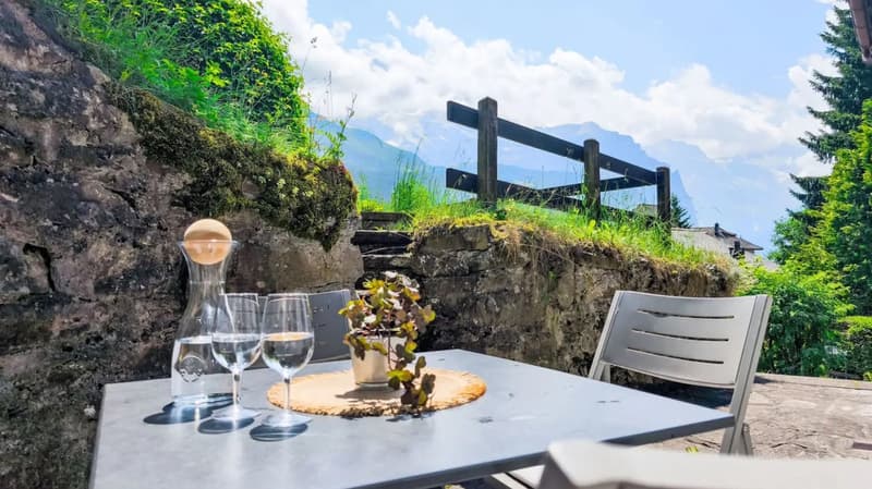 Outdoor dining table with wine glasses, a vase with flowers, and a scenic view of mountains and greenery in the background. The table is set on a stone terrace with a wooden fence overlooking the natural landscape.
