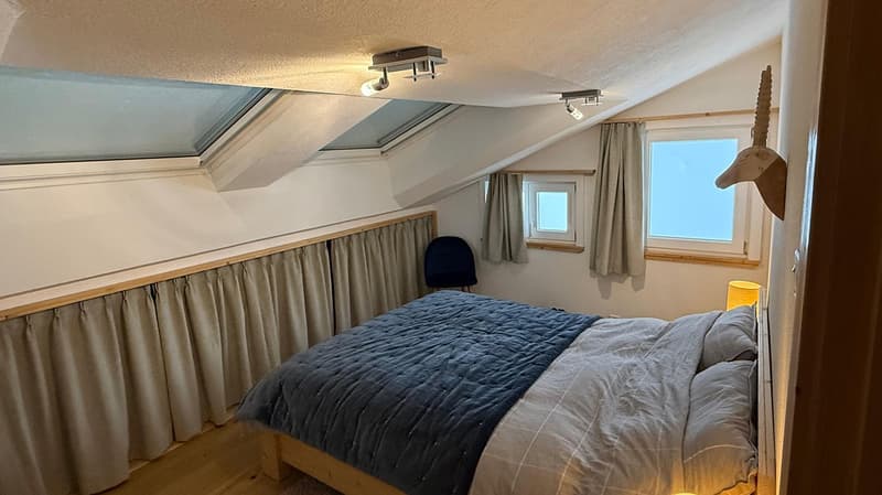 wooden bed, white walls, wooden ceiling, window with curtain, two skylight windows, wooden chair