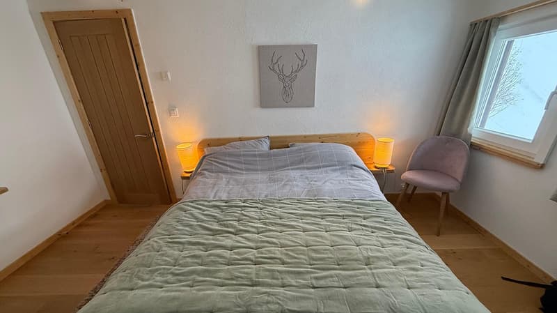bedroom with bed, bedside lamps, chair, deer painting, wooden door, wooden floor, window with curtain