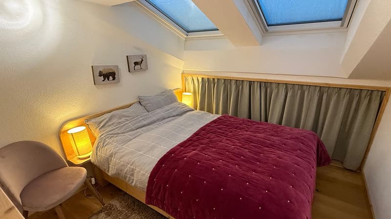Bedroom with wooden bed, nightstand with lamps, carpet, chair, two paintings, two skylights, and curtained window.