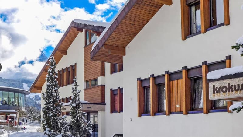 2 story building, white walls, wooden trim, snow on the roof, pine trees, sign reading 'krokus'
