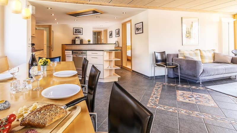 Dining area with a long table, chairs, and a kitchen in the background.