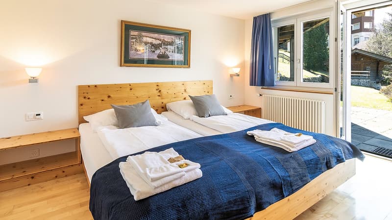 Two twin beds, wooden headboard, white linens, blue blanket, white towels, wooden nightstands, picture frame, wall lamps, white radiator, sliding glass door, outdoor view