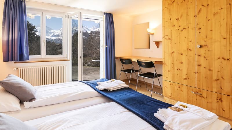Two single beds, wooden wardrobe, desk with two chairs, window with view of mountains, radiator