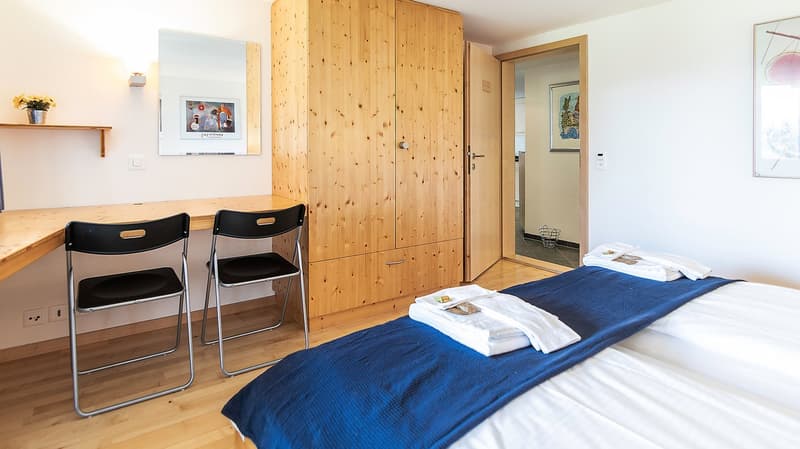 bedroom with desk, two chairs, wooden wardrobe, mirror, bed with blue blanket, white towels