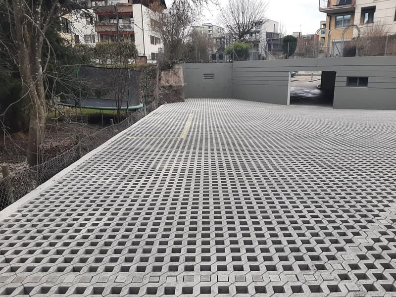 The image shows a paved outdoor area with a grid-like pattern of concrete or stone tiles. There appears to be a trampoline in the background, suggesting this may be a shared outdoor space or courtyard for a residential building. The surrounding buildings