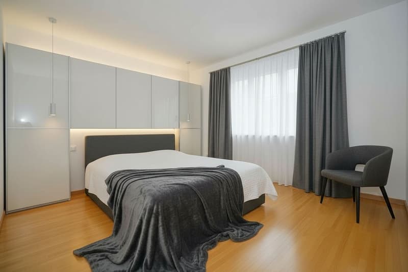 Master bedroom with bed, chair, and window. Wooden floor, gray bed, white bedspread, gray throw blanket. Two lamps hanging from the ceiling. White cabinets with glass doors.