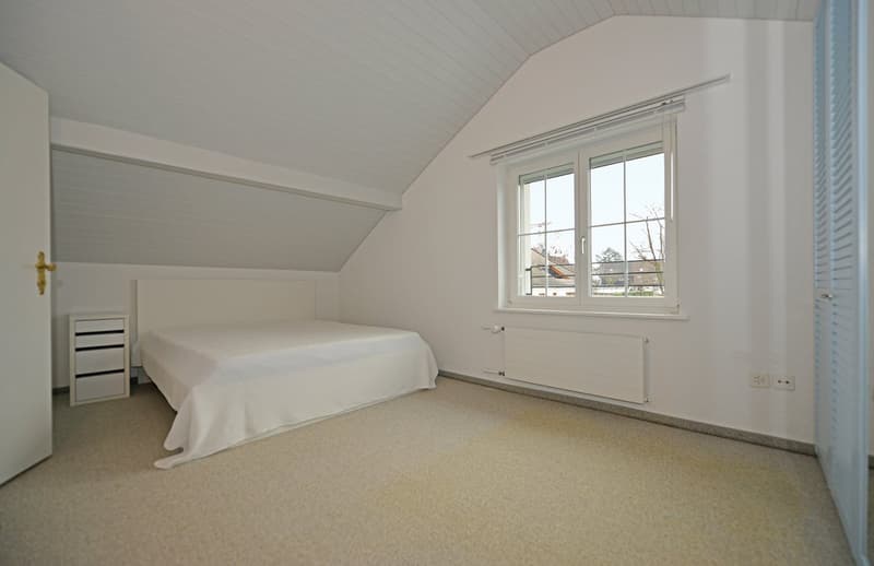 Empty bedroom with white bed, white nightstand, large window, slanted ceiling, radiator.